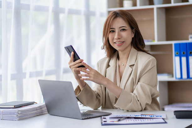 迷人的亚洲女商人在家庭办公室用手机交谈，同时分析每月的财务会计概念笔记本电脑计划。图片下载