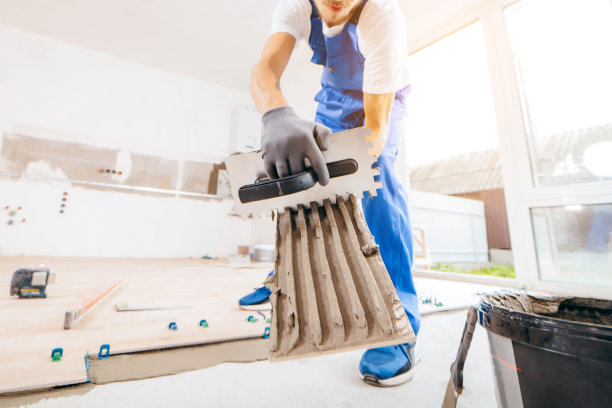 戴着灰色手套、穿着特制制服的修理工正在新房子的地板上铺瓷砖图片下载