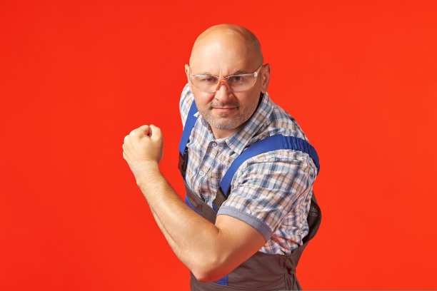 一个穿着工作服、戴着护目镜的秃头男子在红色背景下展示肌肉。图片下载