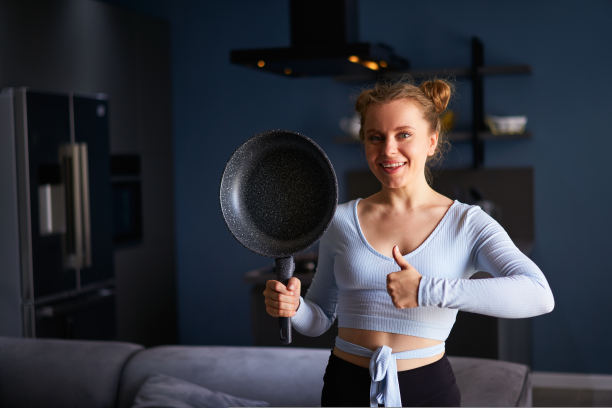 年轻的白人女性微笑的牙齿拿着煎锅与拇指站在家里时髦的现代厨房背景。一个肩上扛着煎锅的快乐的家庭主妇图片下载