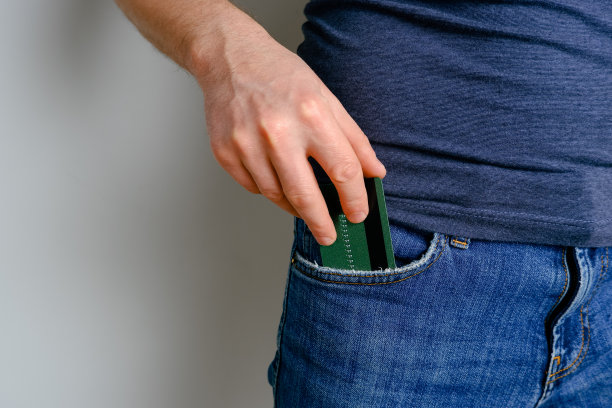 一个年轻人从他的蓝色牛仔裤口袋里拿出一张信用卡。特写一个男人的手从裤子里拿出一张支付卡，背景是浅色的。图片下载