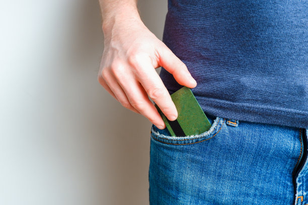 一个年轻人从他的蓝色牛仔裤口袋里拿出一张信用卡。特写一个男人的手从裤子里拿出一张支付卡，背景是浅色的。图片下载