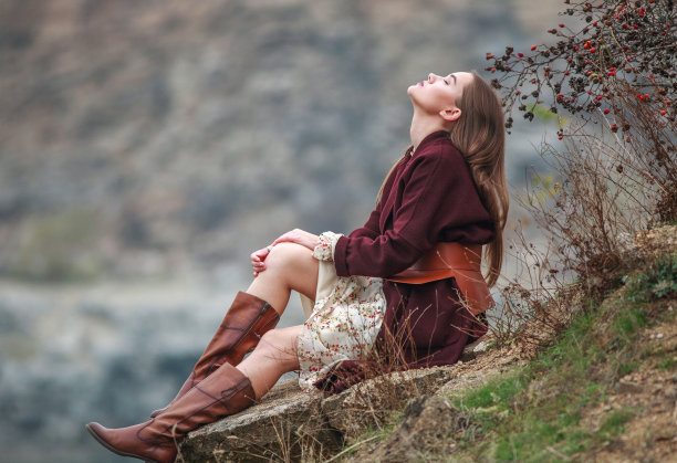 年轻的女士穿着长裙和棕色的靴子坐在石头上，在秋天乡村的岩石和湖的背景上，时尚的时髦女人图片下载