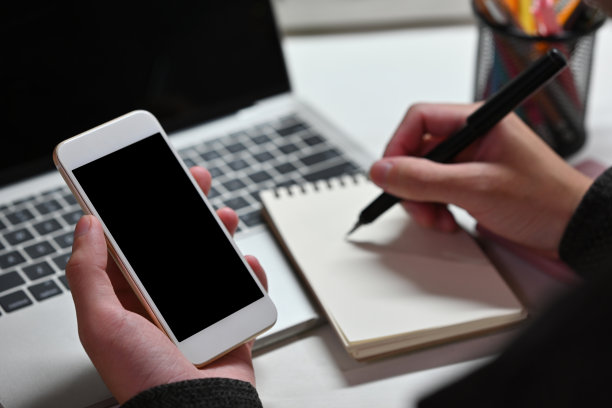 一个男人坐在办公室里用笔记本电脑工作，用智能手机做笔记的特写。为商业、科技、家居概念。图片下载