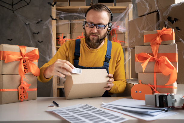 小企业主为万圣节发货包装一个盒子，自由职业者在家庭办公室使用耳机接受网上订单，中小企业，电话营销，万圣节销售图片下载