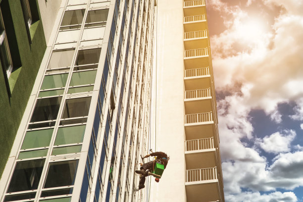 绳子连接工人在房子的墙上。穿着制服的工业登山工人悬挂在蓝天背景下的居民楼上，清洗外墙。工业城市工作。复制文本空间图片下载