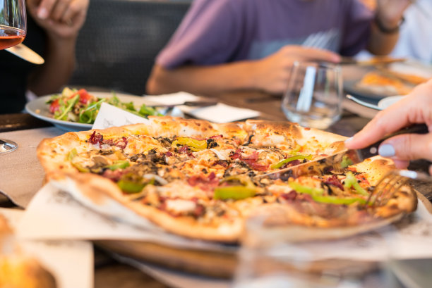 披萨在桌子上，服务的焦点在前景水平照片一个女人正在切一片披萨，人们是背景玫瑰酒是背景图片下载