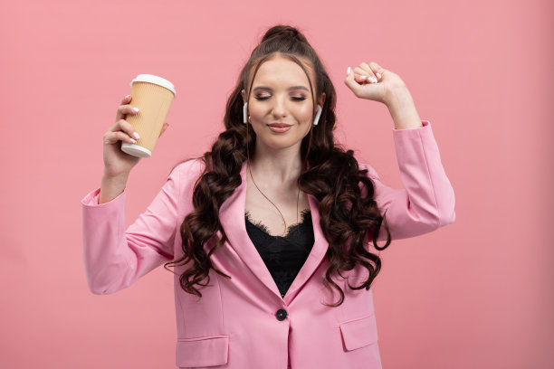 肖像一个穿着优雅的女人在夹克拿着一个外卖杯无线耳机女孩跳舞快乐的粉红色的工作室背景图片下载