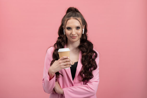一个漂亮的女人用现代科技拿着外卖咖啡，戴着耳机听着广播播客音乐，穿着粉色背景的外套在工作室里图片下载
