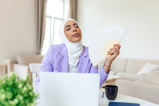 阿拉伯语办公室员工忍受炎热、闷热的空气，夏天在没有空调的家里用笔记本电脑工作。穆斯林商业女性在工作场所用手持风扇降温图片下载