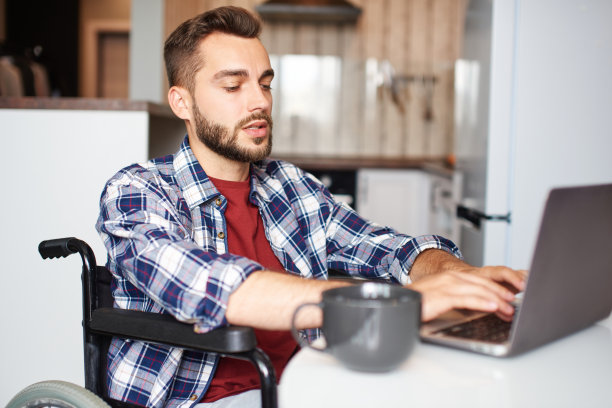 年轻英俊的男子坐在轮椅上在家里用笔记本电脑工作图片下载