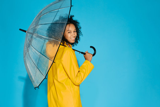 身穿黄色防水雨衣的非裔美国妇女，伞是透明的，被隔离在蓝色的工作室背景上。秋季的概念图片下载
