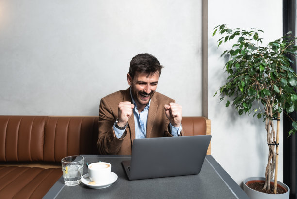 年轻的成功商人坐在自助餐厅喝咖啡，看起来很高兴股票市场价格上涨。男性在他的新业务中致富，在网上交易和财务方面取得了成功。图片下载