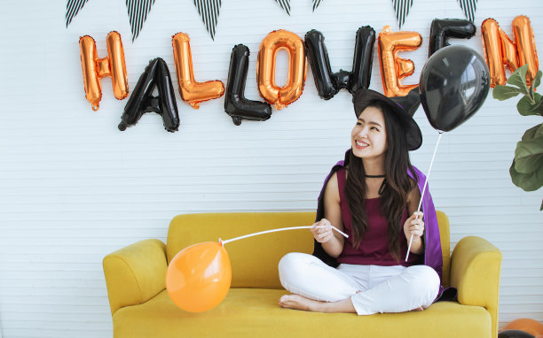 画像亚洲美丽的女子穿着女巫的服装装饰和庆祝万圣节的聚会，微笑着快乐和乐趣，玩气球，坐在家里的客厅。图片下载