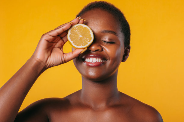非裔美国妇女，带着自信的微笑，用一片隔离在橙色背景上的柠檬堵住一只眼睛。美丽女人的身体。柑橘卫生保健。图片下载