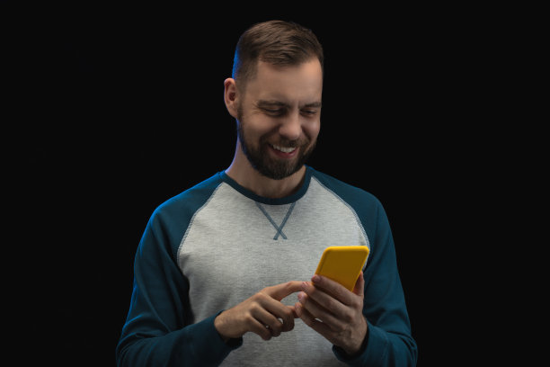 工作室肖像的微笑的人打赌最喜欢的球队在博彩公司的网站使用智能手机，孤立的黑色背景。体育投注、赌博、金钱赢的概念图片下载