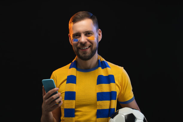 工作室肖像微笑的幸运的足球球迷男子拿着球在他的手，在博彩公司的网站上使用智能手机下注最喜欢的球队，孤立在黑色背景图片下载