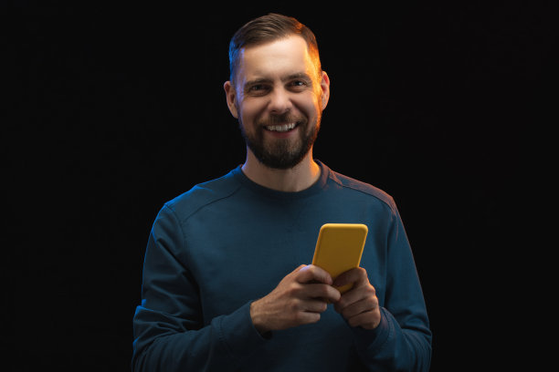 工作室的肖像英俊的微笑，穿着随意的胡子男子摆姿势与手机在黑色背景上孤立的手图片下载