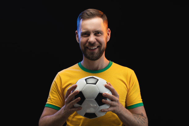 工作室的形象英俊的微笑英俊的大胡子的家伙在黄色t恤是摆姿势在黑色背景与足球在手中图片下载