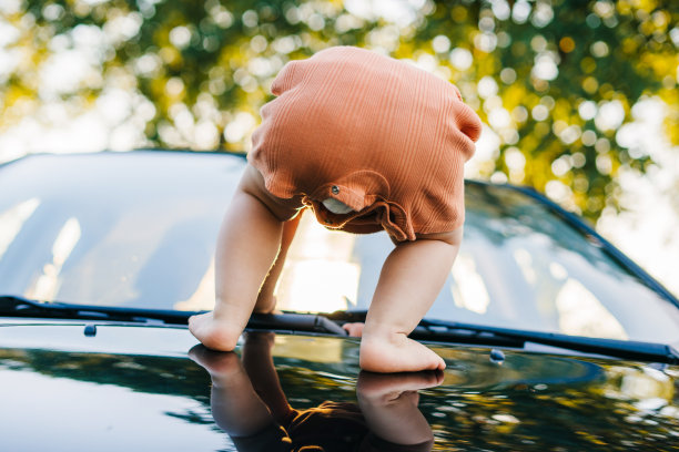 婴儿试图站在停在路边的汽车的引擎盖上。暑假的乐趣。年轻漂亮的女孩。幸福的家庭。图片下载