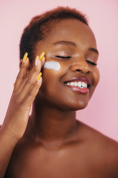 美丽的非裔美国妇女在她的脸颊上涂抹保湿霜，隔离在粉红色的背景上。抗衰老护肤品。美女面部皮肤护理图片下载