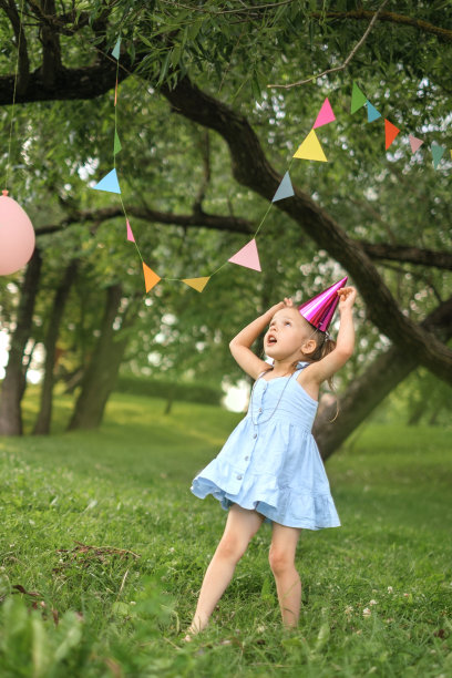 一个孩子戴着节日的帽子，穿着蓝色的衣服，在花园里玩挂在树上的花环，庆祝他的生日。图片下载