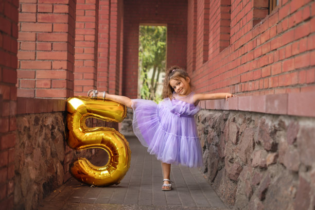一个5岁的体操女孩穿着紫色的节日服装，手里拿着数字形状的金色气球，摆出芭蕾舞演员的姿势。孩子庆祝一周年图片下载