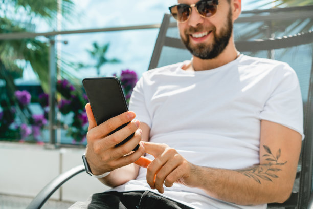 拍摄的快乐微笑着英俊的年轻人浏览他的智能手机，躺在甲板椅上冷却。图片下载