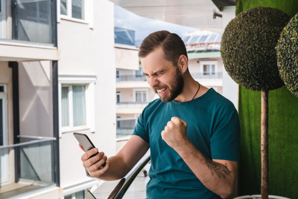 年轻的穿着随意的英俊的大胡子男子在他的手机上阅读信息，并作出胜利者的手势高兴地得到好消息图片下载