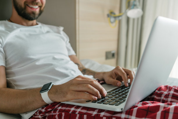 裁剪拍摄的快乐微笑的年轻人穿着睡衣是在他的笔记本电脑上工作，而坐在他的床上图片下载