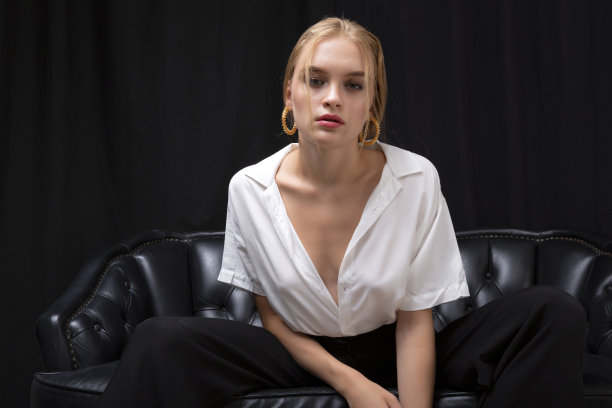 身穿白衬衫和黑色布裤的金发女郎坐在黑色皮沙发上，背景是黑色窗帘。图片下载