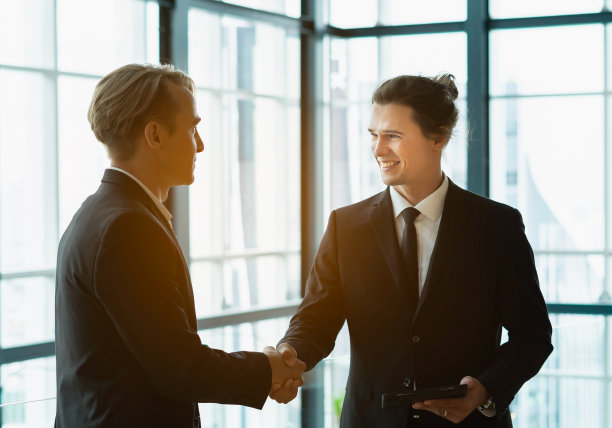 商人与伙伴握手，微笑着看着男人。同事在现代办公室工作，有窗口背景图片下载