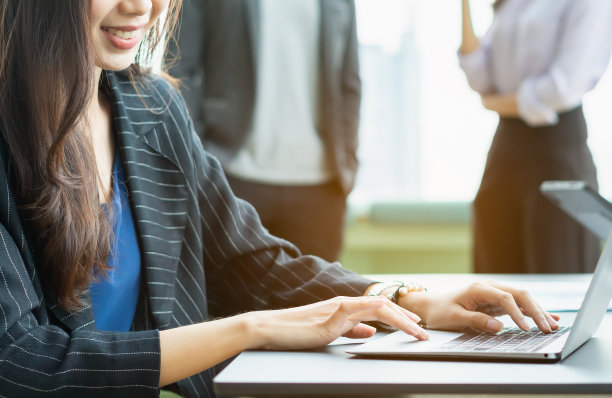 近距离的手，女商人坐着，微笑，和笔记本电脑工作。在轻松的工作场所，一群人站在窗口背景下讨论图片下载