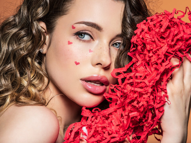 富有表现力和感性的模特与明亮的妆容，持有明亮的彩纸近脸。画的是一个美丽的女孩，脸上画着红心。时髦的时尚照片与白人模特。图片下载