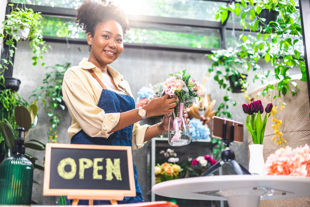 非裔美国女性影响力或中小企业所有者微笑工作在家庭摄像机自拍。面带微笑的女花商打电话。年轻女子在花店工作与开放的标志..图片下载