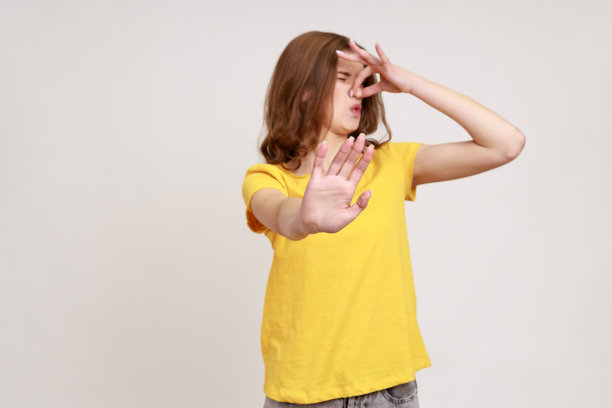 身穿黄色t恤的不满少女的肖像，厌恶地扮鬼脸，用手指捏鼻子以避免难闻的气味，并做出停止的手势。图片下载