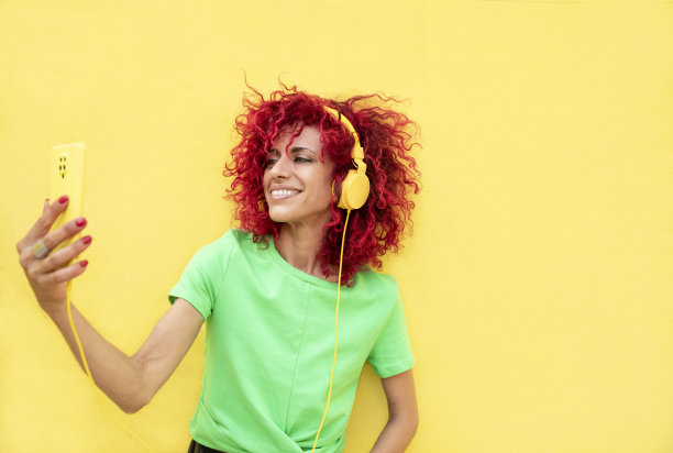 横向肖像的拉丁妇女与非洲红头发拿着手机，享受听音乐与黄色耳机在孤立的黄色背景图片下载