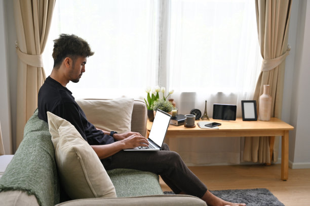穿着休闲装的卷发帅哥在客厅用笔记本电脑，笔记本电脑的空白屏幕，家庭生活和家庭的概念。图片下载