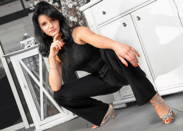 年轻的褐发女子在黑色衬衫和裤子是蹲在高跟鞋，用她的手指支撑她的下巴，看着相机背景的白色衣柜的抽屉。图片下载