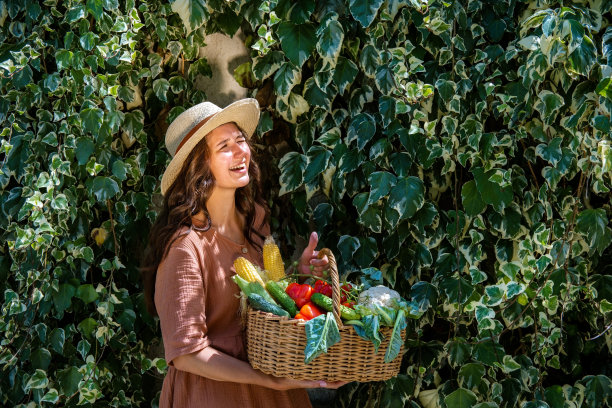 美丽的白人妇女戴草帽和亚麻衣服在花园里与篮子蔬菜作物在常春藤覆盖的花园墙。图片下载