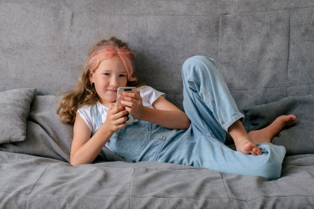 小女孩躺在沙发上玩智能手机。儿童电子产品成瘾的概念。图片下载