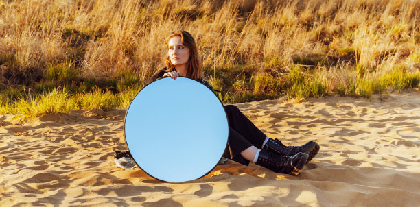 户外大气的生活方式的照片年轻美丽的黑发女人在黑色的商务套装坐在海滩与天空反射在镜子。带镜子的概念性照片。图片下载