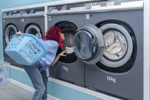 一个留着红色爆炸头的年轻拉丁妇女在蓝色自动洗衣店的工业洗衣机里找袜子图片下载