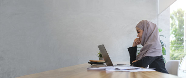 侧面看戴头巾的女商人坐在现代工作场所，使用笔记本电脑。复制空间为您的广告文本。图片下载