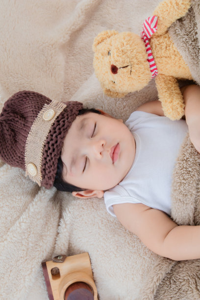亚洲可爱的新生儿戴着棕色针织帽，深睡在米色毯子上，旁边是泰迪熊和玩具相机，安全舒适。小幼童吃饱后可爱的休息。图片下载