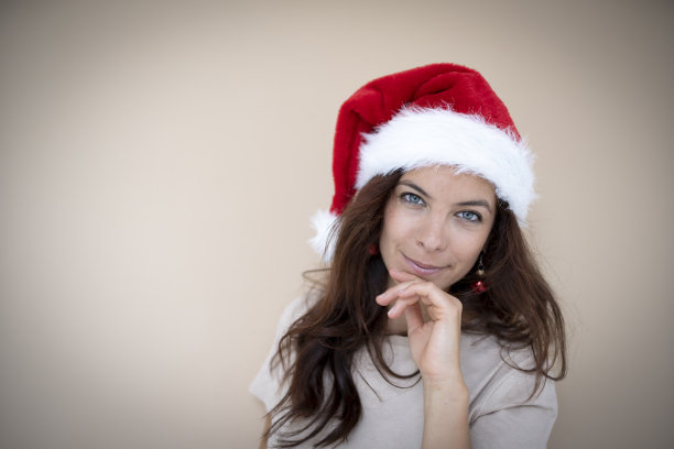 漂亮的年轻女子站在棕色背景前，戴着圣诞帽和红色耳环，形状像圣诞树球图片下载