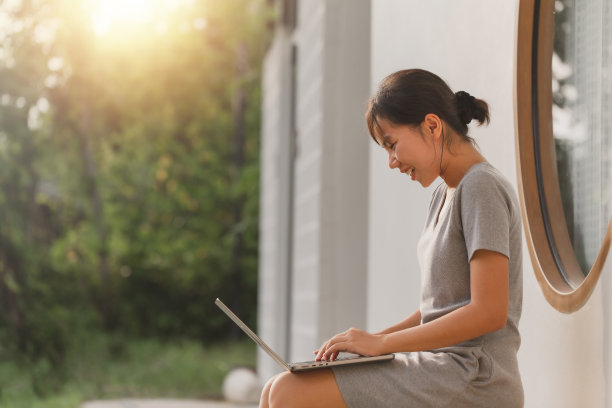 年轻有吸引力的亚洲自由职业女性坐在窗前的房子外面的长凳上工作的笔记本电脑在暑假。快乐亚洲青少年学生在线学习图片下载