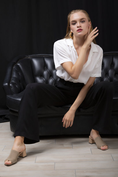 身穿白衬衫和黑色布裤的金发女郎坐在黑色皮沙发上，背景是黑色窗帘。图片下载