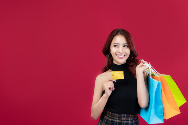 时尚的亚洲女人在黑色的衣服拿着购物袋，在深红色的背景下持有黄金信用卡。美丽的泰国妇女拿着购物袋，里面有深红色背景的金色信用卡。图片下载