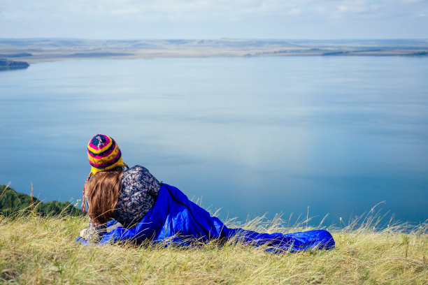 女旅行者裹在蓝色睡袋上的草湖背景山图片下载